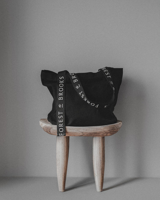 Black tote bag with 'Forest & Brooks' branding on a wooden stool against a gray background